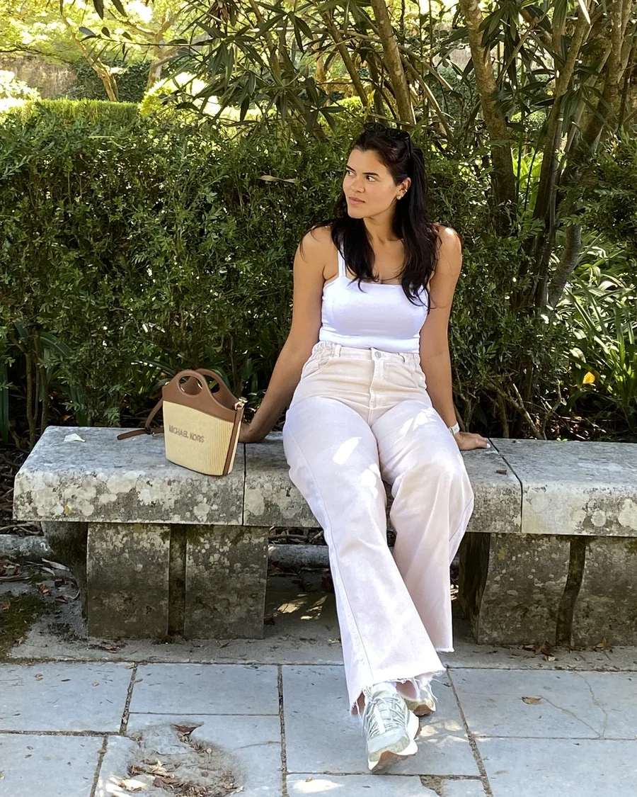 Talitha sentada num banco de pedra em um jardim, de blusa branca e calça clara, olhando para o lado, com uma bolsa de palha ao lado.